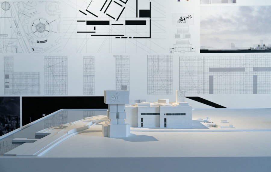 An architectural model on a table in front of pinned-up drawings